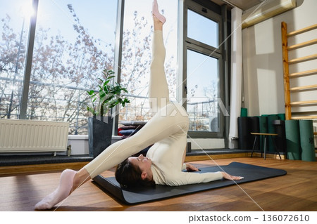 Woman practicing advanced plow pose variation in yoga studio 136072610