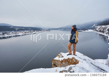 Woman tourist on a snowy hilltop overlooking the Yenisei River and a winter village. Winter adventure travel and exploration concept. 136073184