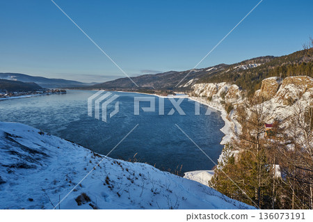 Blue water of the Yenisei River flowing through a winter landscape with snow-covered banks and distant mountains under a clear sky. Blue water of the Yenisei River flowing through a winter landscape with snow-covered banks and distant mountains under a clear sky. 136073191