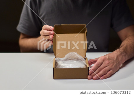 Person unpacks device, Individual reveals compact electronic component inside, Person diligently removes protective cover to access electronic device within home environment 136073312