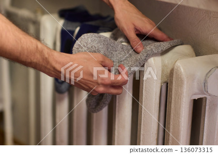 Person placing moist wool socks onto heated radiator fins, Detailed scene of hand carefully positioning damp wool socks on heated metal radiator with knit texture 136073315