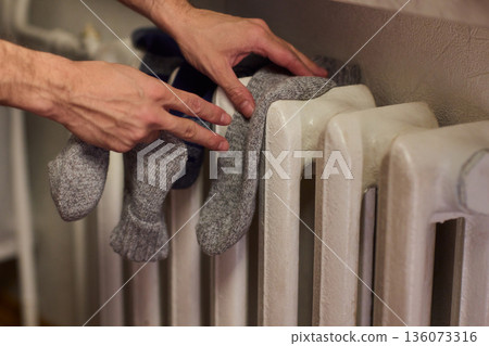 Person placing moist wool socks onto heated radiator fins, Detailed scene of hand carefully positioning damp wool socks on heated metal radiator with knit texture Person placing moist wool socks onto heated radiator fins, Detailed scene of hand carefully positioning damp wool socks on heated metal radiator with knit texture 136073316