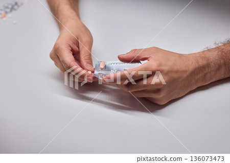Hands extracting capsule from blister pack gently, closeup on fingertips and foil on white tabletop, quiet moment of dosing accuracy and calm preparation for intake Hands extracting capsule from blister pack gently, closeup on fingertips and foil on white tabletop, quiet moment of dosing accuracy and calm preparation for intake 136073473