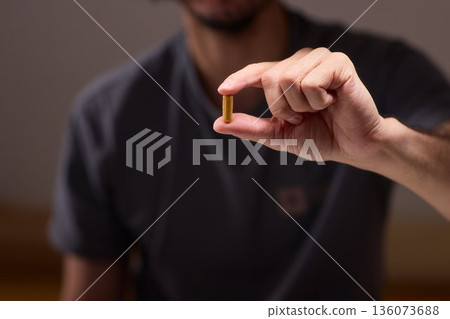 Person carefully holding pill amid scattered tablets and bottles, Individual attentively grasping single pill among various tablets and bottles in warm ambient light 136073688