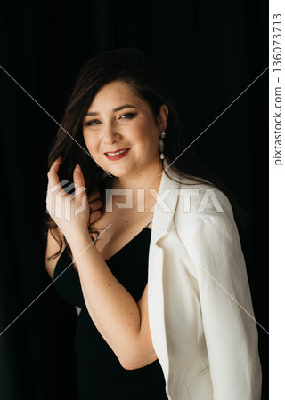 Beautiful smiling brunette posing fixing her long, well-groomed hair. Portrait of a woman with dark hair. 136073713
