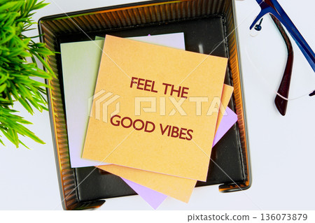 A stack of colorful sticky notes rests in a black organizer tray on a white desk. The top note displays Feel the good vibes in bold red letters, beside a green plant and blue glasses 136073879