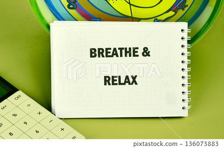 A white spiral bound notepad sits on a smooth green surface, displaying the bold words BREATHE and RELAX. Part of a calculator and a colorful, circular object are visible in the frame 136073883