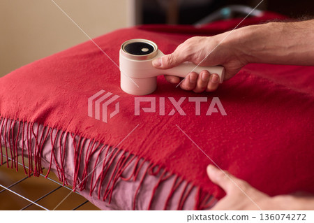 Hands cleaning red wool scarf with lint roller across fringed fabric, closeup texture, warm bedroom lighting, careful motion, pet hair removal, garment ready for outing 136074272