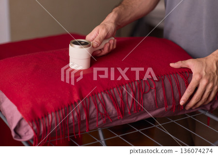 Hands cleaning red wool scarf with lint roller across fringed fabric, closeup texture, warm bedroom lighting, careful motion, pet hair removal, garment ready for outing 136074274