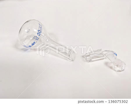 A shattered Erlenmeyer flask and its broken pieces are shown on a white background, symbolizing accidents in the lab 136075302