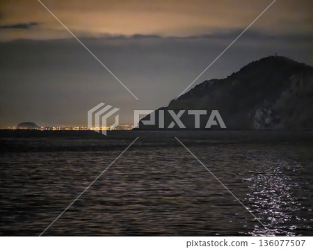 Nighttime coastal vantage showcases glowing city reflections and tranquil yet alert atmosphere 136077507