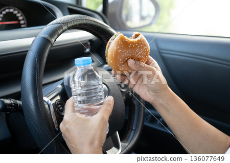 Asian woman driver hold and eat hamburger and french fries in car, dangerous and risk an accident. 136077649