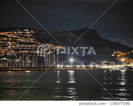 Night marina with rows of illuminated masts and calm water reflecting amber lights, bustling harbor energy Night marina with rows of illuminated masts and calm water reflecting amber lights, bustling harbor energy 136077896
