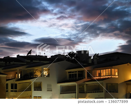 Evening glow over peaceful seaside homes, Calm dusk ambiance with illuminated balconies and gentle breezes Evening glow over peaceful seaside homes, Calm dusk ambiance with illuminated balconies and gentle breezes 136077899