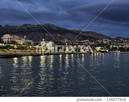 Waterfront evening scene with glowing city lights, City skyline reflects brightly on rippling water at dusk Waterfront evening scene with glowing city lights, City skyline reflects brightly on rippling water at dusk 136077926