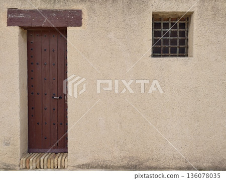 Minimalist composition highlighting tall wooden door beside barred window on muted plaster facade Minimalist composition highlighting tall wooden door beside barred window on muted plaster facade 136078035