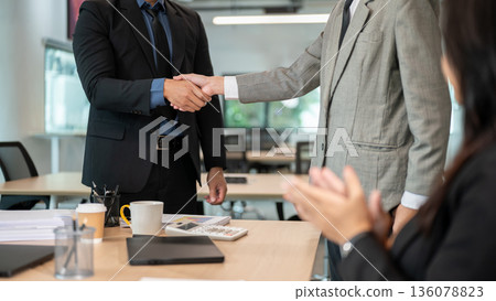 Businessman standing and shaking hands while woman coworker clapping aside at wooden table in office Businessman standing and shaking hands while woman coworker clapping aside at wooden table in office 136078823