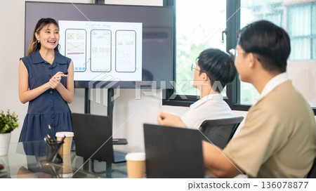 Asian woman presenting mobile app design on monitor display screen to coworkers in an office meeting 136078877