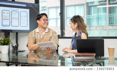 Asian glasses man talking while holding and showing tablet to woman coworker in office meeting room. Asian glasses man talking while holding and showing tablet to woman coworker in office meeting room. 136078903