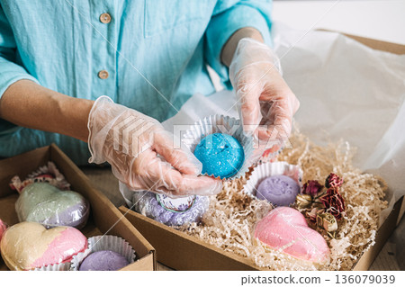 hands place blue bath bomb into cardboard gift box filled with shredded paper and various colorful cosmetic products. ceramide bath bomb, probiotic soak, lipid repair, skin longevity. hands place blue bath bomb into cardboard gift box filled with shredded paper and various colorful cosmetic products. ceramide bath bomb, probiotic soak, lipid repair, skin longevity. 136079039