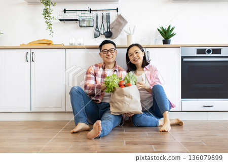 A smiling Asian couple sits on the floor with a bag of fresh produce in their modern kitchen 136079899