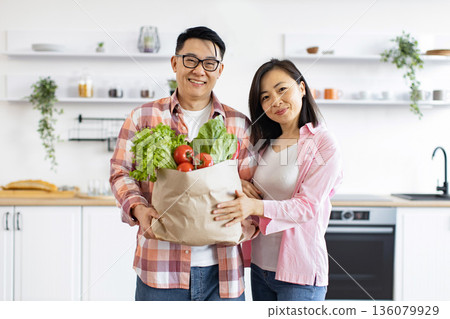 An Asian couple smiles while holding a bag of fresh produce in their modern kitchen 136079929