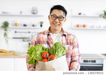 An Asian man smiles while holding a bag of fresh produce in his kitchen An Asian man smiles while holding a bag of fresh produce in his kitchen 136079932