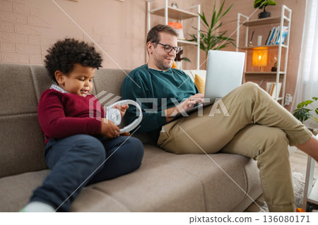 Father working on laptop at home next to his son 136080171