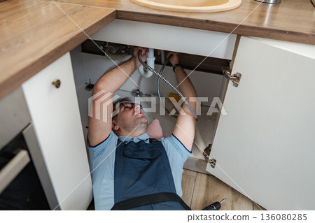 Handyman repairing kitchen sink drain pipe below cabinets 136080285