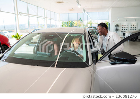 Saleswoman helping customer sitting inside new car at dealership 136081270