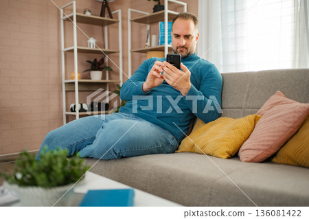 Man relaxing on sofa using smartphone for internet browsing Man relaxing on sofa using smartphone for internet browsing 136081422