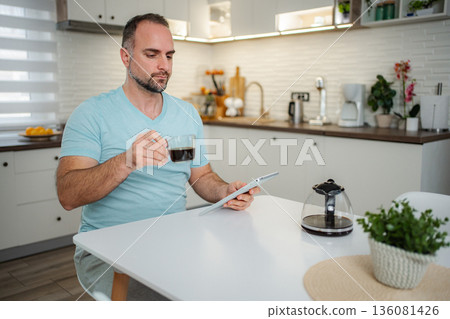 Adult man drinking coffee and using tablet in kitchen Adult man drinking coffee and using tablet in kitchen 136081426