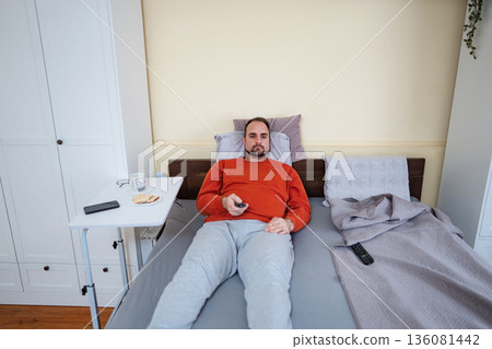 Man relaxing in bed watching television with remote 136081442