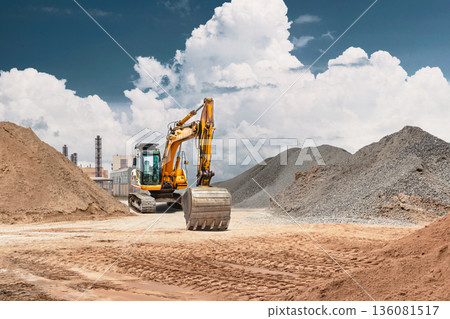 A powerful excavator with a large bucket at a construction site surrounded by piles of gravel and sand, under a blue sky with clouds 136081517