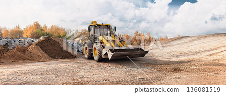 A loader operates on a construction site, moving dirt while surrounded by piles of earth and rocky materials under a cloudy sky in autumn A loader operates on a construction site, moving dirt while surrounded by piles of earth and rocky materials under a cloudy sky in autumn 136081519
