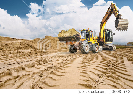 Heavy machinery moves through sandy ground making deep tracks while a loader and excavator work in the background on a cloudy day at a construction site 136081520
