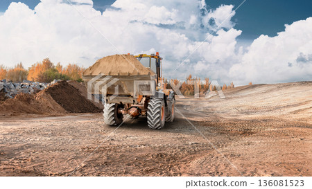 A loader is moving dirt on a construction site during daytime. The scene shows an open area with earth moved and clouds in the sky A loader is moving dirt on a construction site during daytime. The scene shows an open area with earth moved and clouds in the sky 136081523