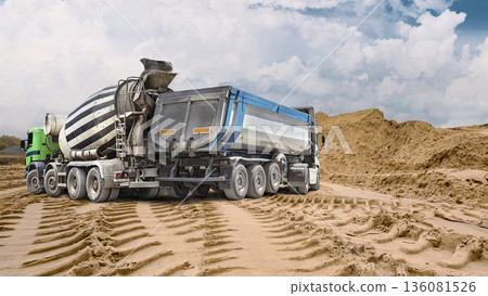 A large dump truck and concrete mixer truck at a construction site on a cloudy day. A large pile of sand is visible in the background 136081526