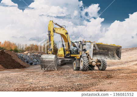 Loader and excavator work together at a construction site to move dirt and prepare the area for upcoming projects in bright daylight Loader and excavator work together at a construction site to move dirt and prepare the area for upcoming projects in bright daylight 136081552