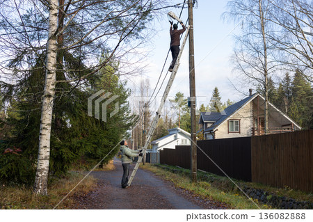 Two adult men using ladder at utility pole when replacing lamp on rural road surrounded by trees and houses during autumn daylight. Two adult men using ladder at utility pole when replacing lamp on rural road surrounded by trees and houses during autumn daylight. 136082888