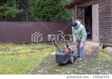 Man checks lawnmower functionality, dressed warmly, beside log cabin grassy area cool weather conditions in overcast day. Man checks lawnmower functionality, dressed warmly, beside log cabin grassy area cool weather conditions in overcast day. 136082919