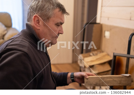 Senior male loading wooden log into stove, lights fireplace inside living. Space winter warmth concept. 136082944