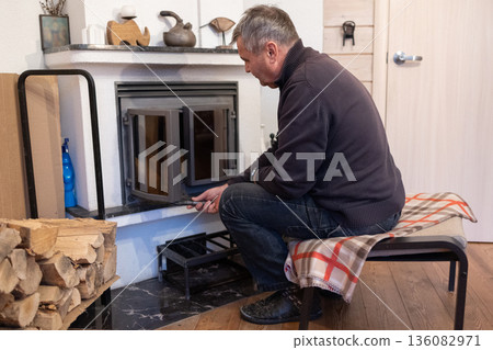 Elderly man lights fireplace into indoor fireplace preparing home heating during cold season domestic routine. 136082971