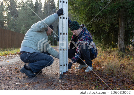Two adult men using ladder at utility pole when replacing lamp on rural road surrounded by trees and houses during autumn daylight. Two adult men using ladder at utility pole when replacing lamp on rural road surrounded by trees and houses during autumn daylight. 136083002