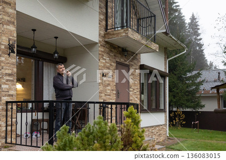 Mature man standing on balcony holding cigarette calm thoughtful mood, casual clothing in residential exterior. Everyday lifestyle moment. Mature man standing on balcony holding cigarette calm thoughtful mood, casual clothing in residential exterior. Everyday lifestyle moment. 136083015