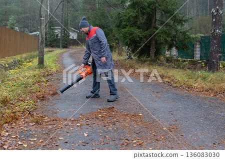 Adult man operating leaf blower on wet asphalt road, autumn season, routine outdoor cleaning, practical work activity in residential setting. 136083030