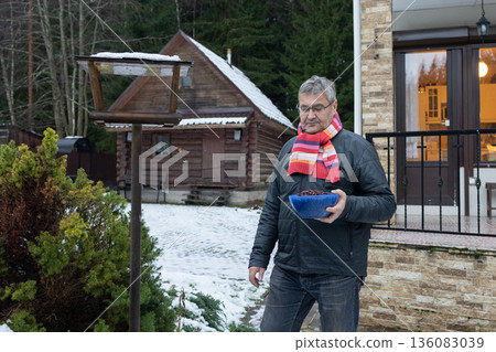 Man holding bowl of seeds while servicing wooden bird feeder outdoors with snow on ground and evergreen plants. 136083039