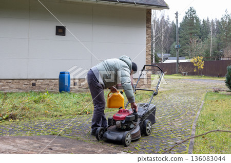 Man in wearing warm jacket and beanie fills lawnmower fuel tank with yellow can near shed. Overcast day outdoor maintenance. 136083044