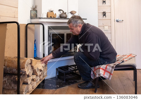 Elderly man lights fireplace into indoor fireplace preparing home heating during cold season domestic routine. Elderly man lights fireplace into indoor fireplace preparing home heating during cold season domestic routine. 136083046