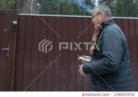Adult man using smartphone smart home application to open residential gate in snowy suburban environment. Adult man using smartphone smart home application to open residential gate in snowy suburban environment. 136083050